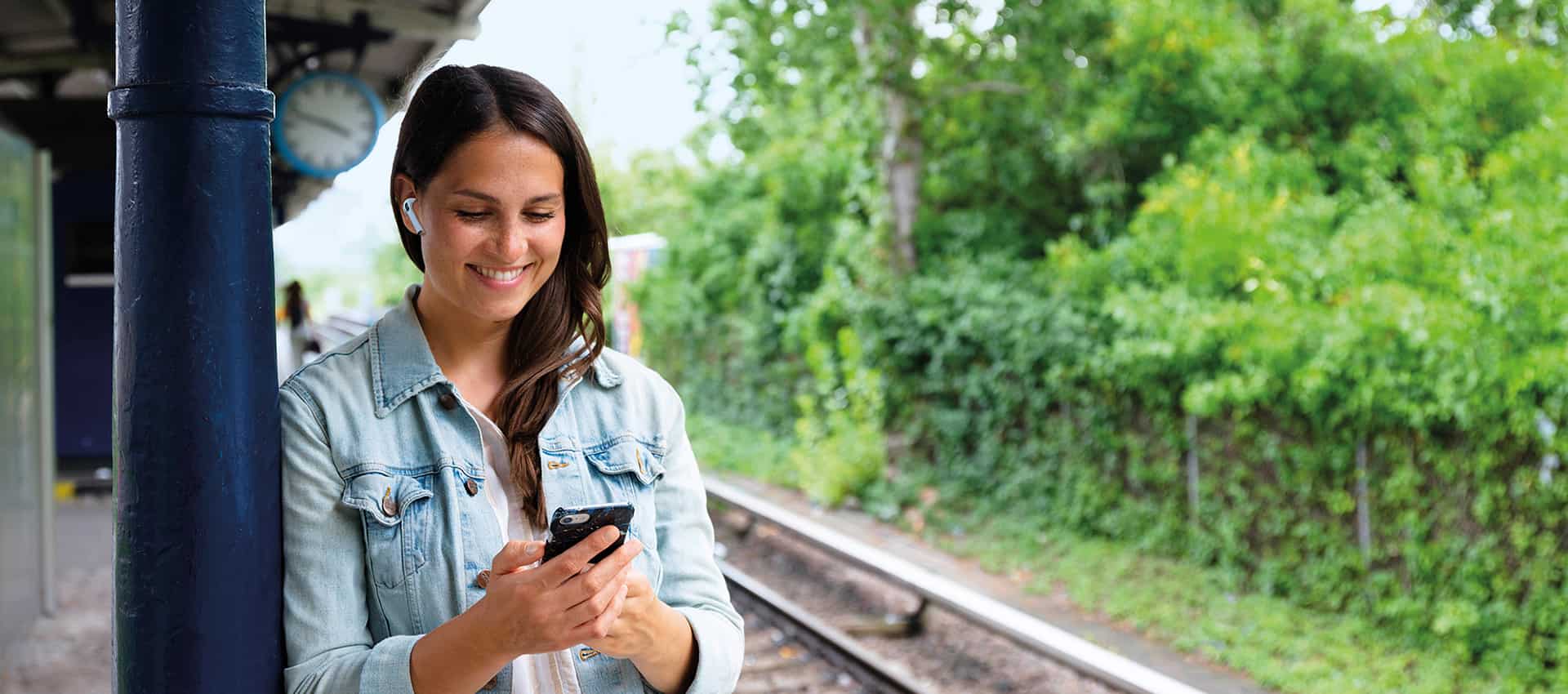 Junge Frau steht auf Bahnsteig und hört mit ihrem Handy ein Hörbuch.